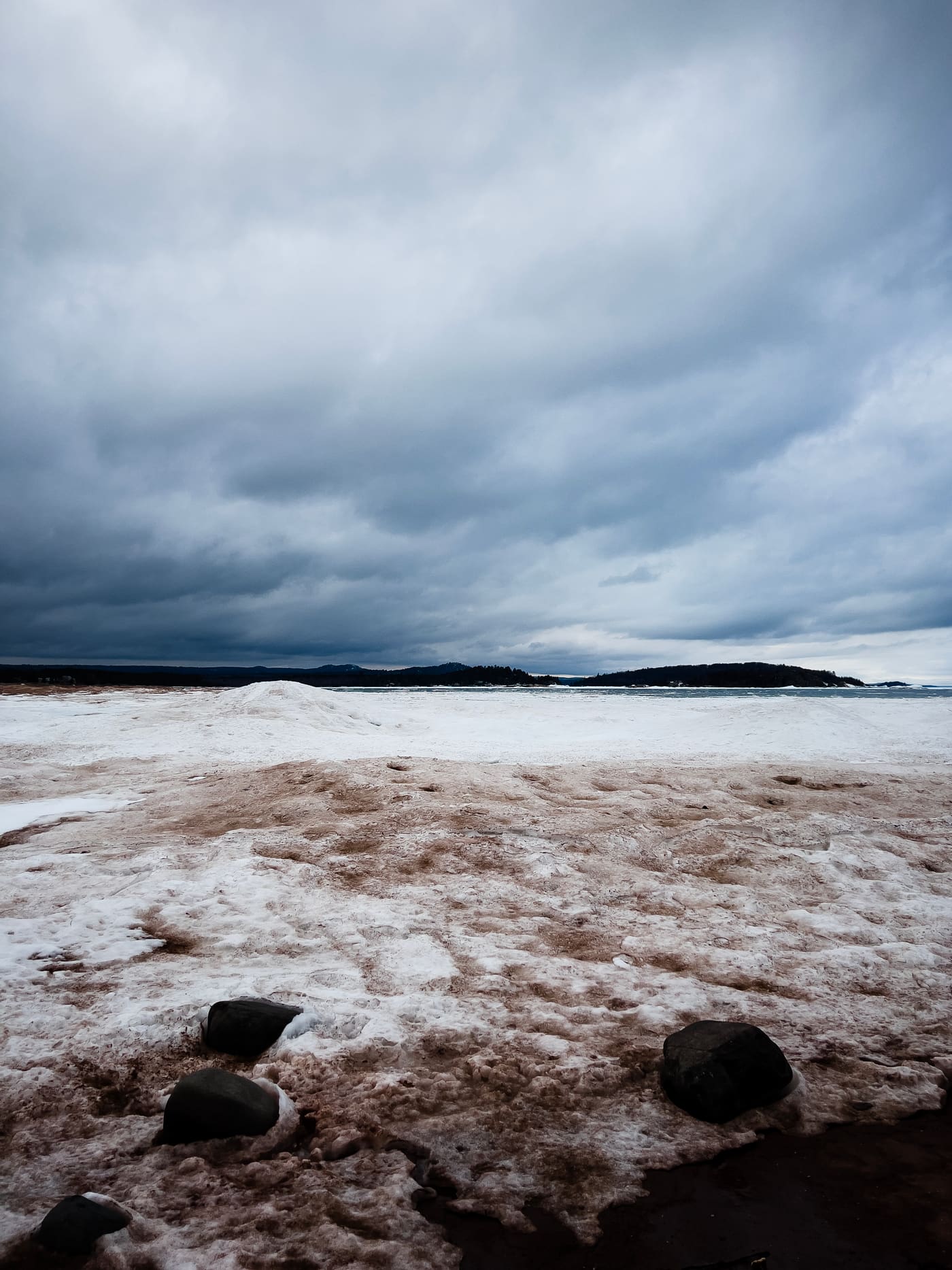 Snow and ice-covered shoreline with dark boulders beneath a vast bank of grey storm clouds over distant headlands.