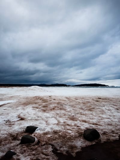 Snow and ice-covered shoreline with dark boulders beneath a vast bank of grey storm clouds over distant headlands.