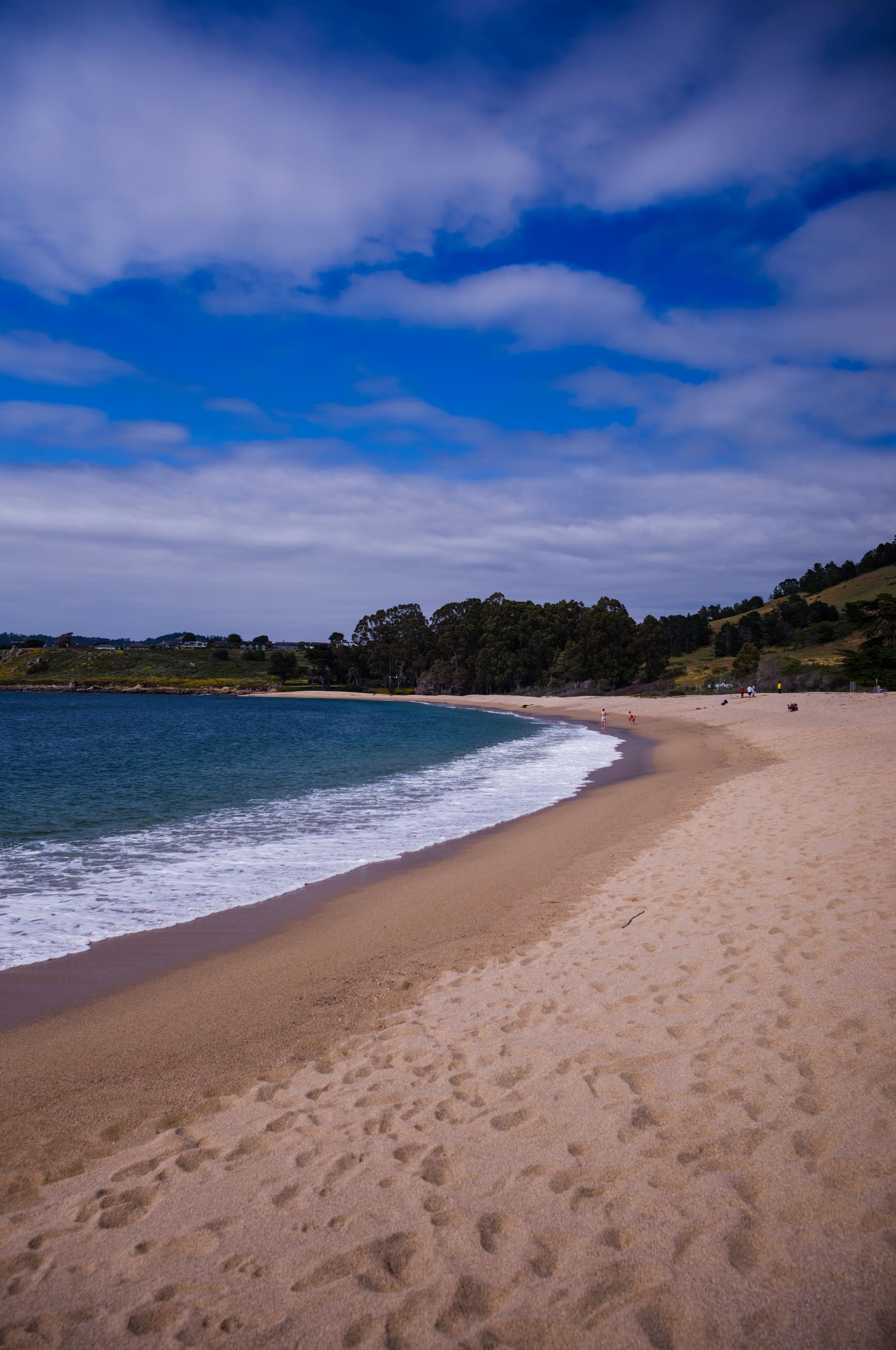 Wide sweep of pale sand curving beside turquoise surf under a dramatic blue sky streaked with white cloud.