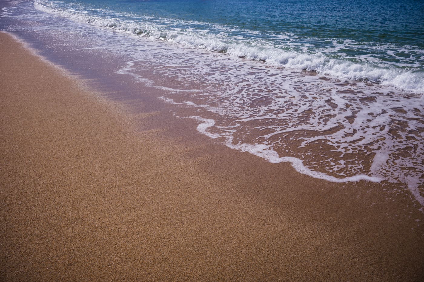 Diagonal shoreline where white sea foam slides over golden wet sand, with turquoise surf breaking beyond.