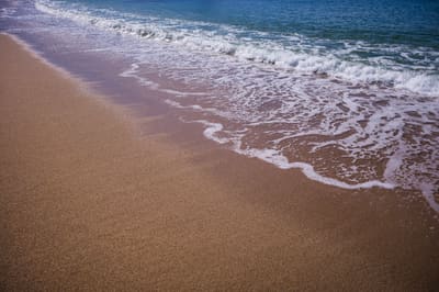 Diagonal shoreline where white sea foam slides over golden wet sand, with turquoise surf breaking beyond.