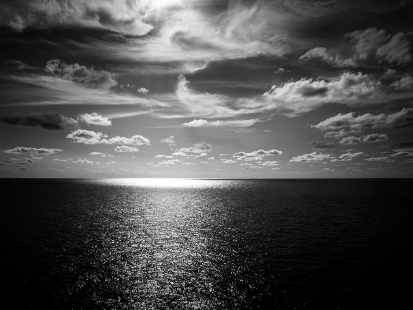 Black and white seascape with sun glare on the horizon casting a bright path across rippled open ocean under scattered clouds.