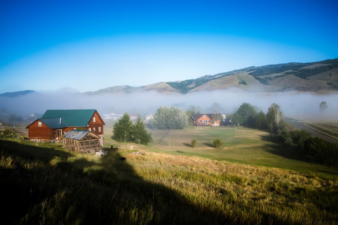 A green-roofed log home and pasture below rolling hills wrapped in low morning fog under deep blue sky.