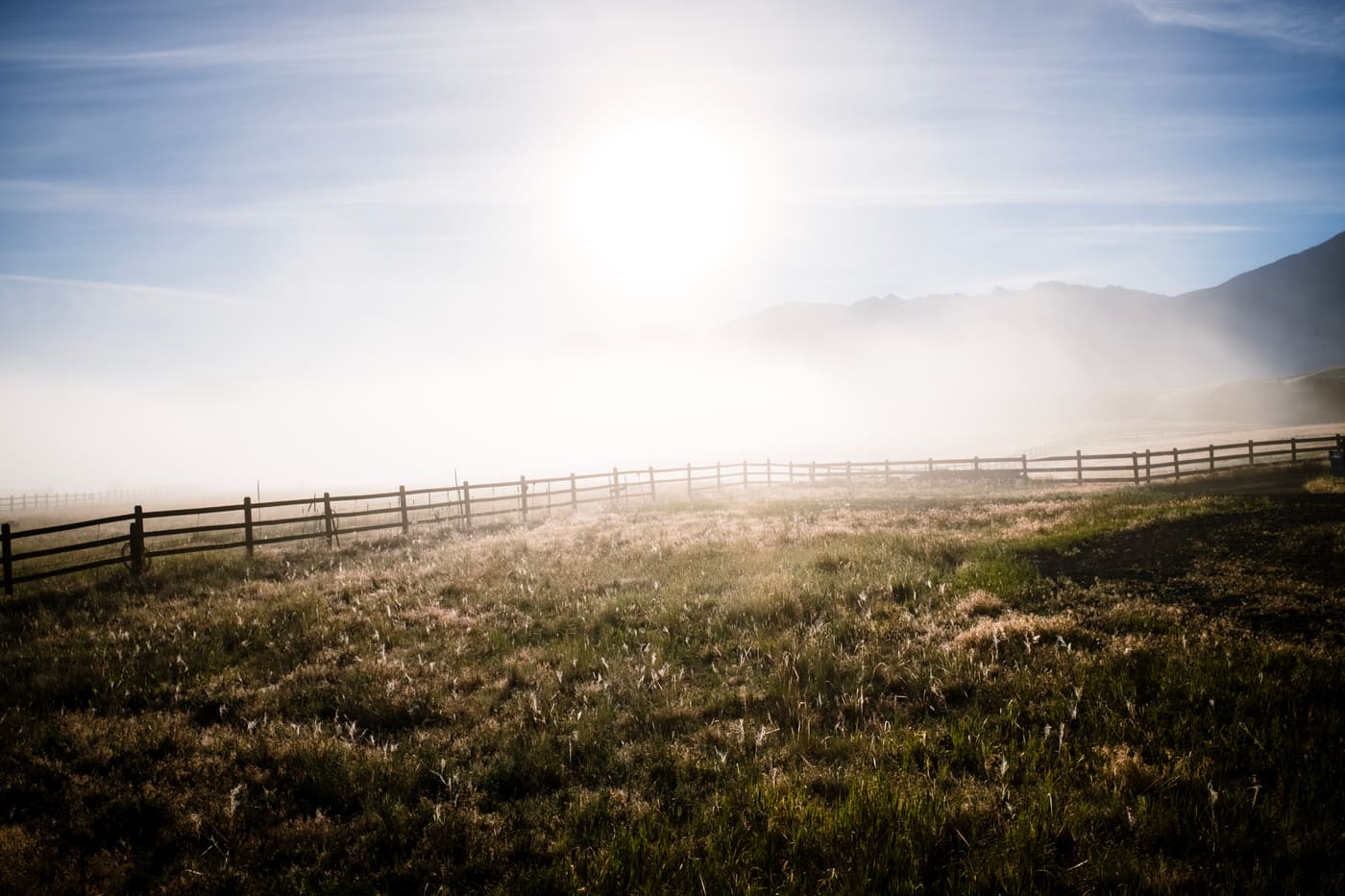 Sun burning through low fog above a wooden post-and-rail fence bordering a dry grass field with distant hills.