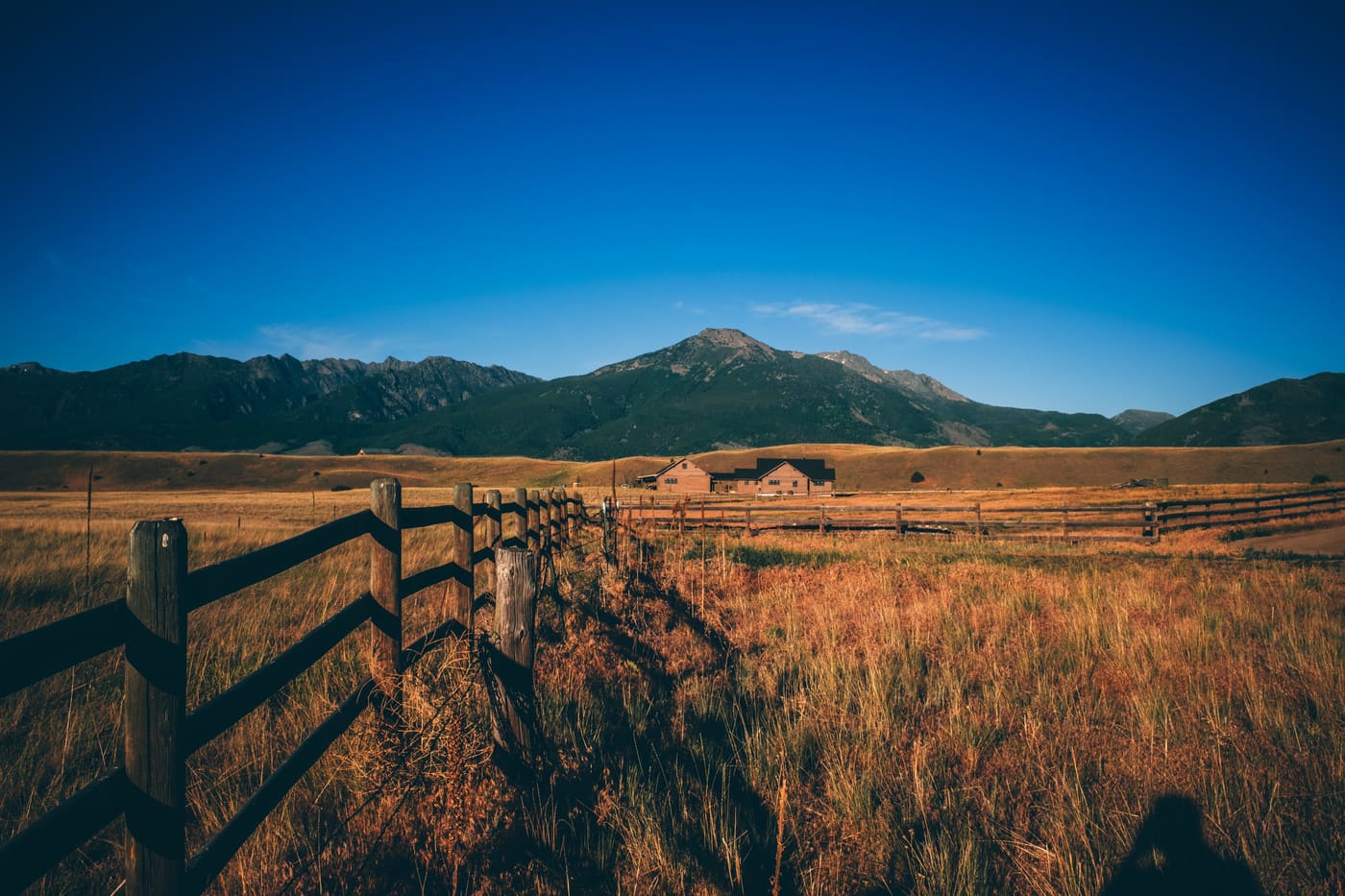 Weathered wooden fence crossing golden ranch grassland toward distant barns and mountains under deep blue sky