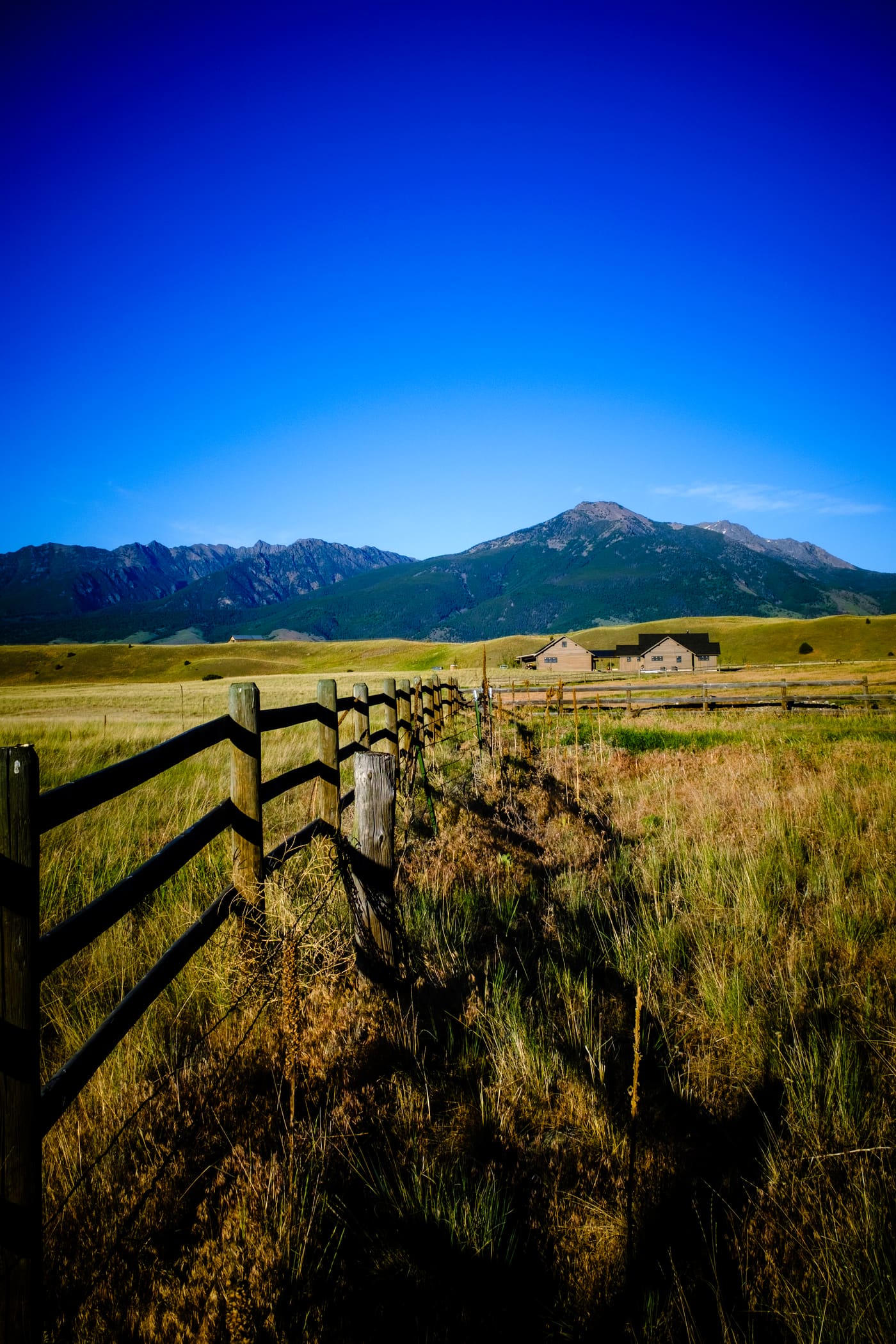 Weathered wooden rail fence leading across a golden grass field toward green mountains under a deep blue sky.