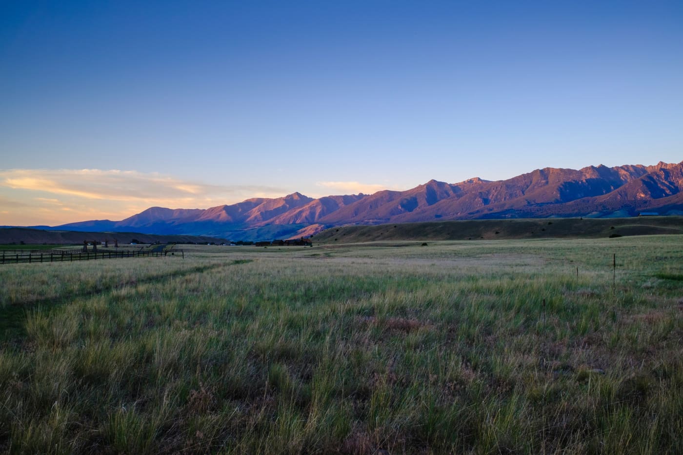 Alpenglow lights a long mountain range above a broad grass valley with a wooden fence line at dusk.