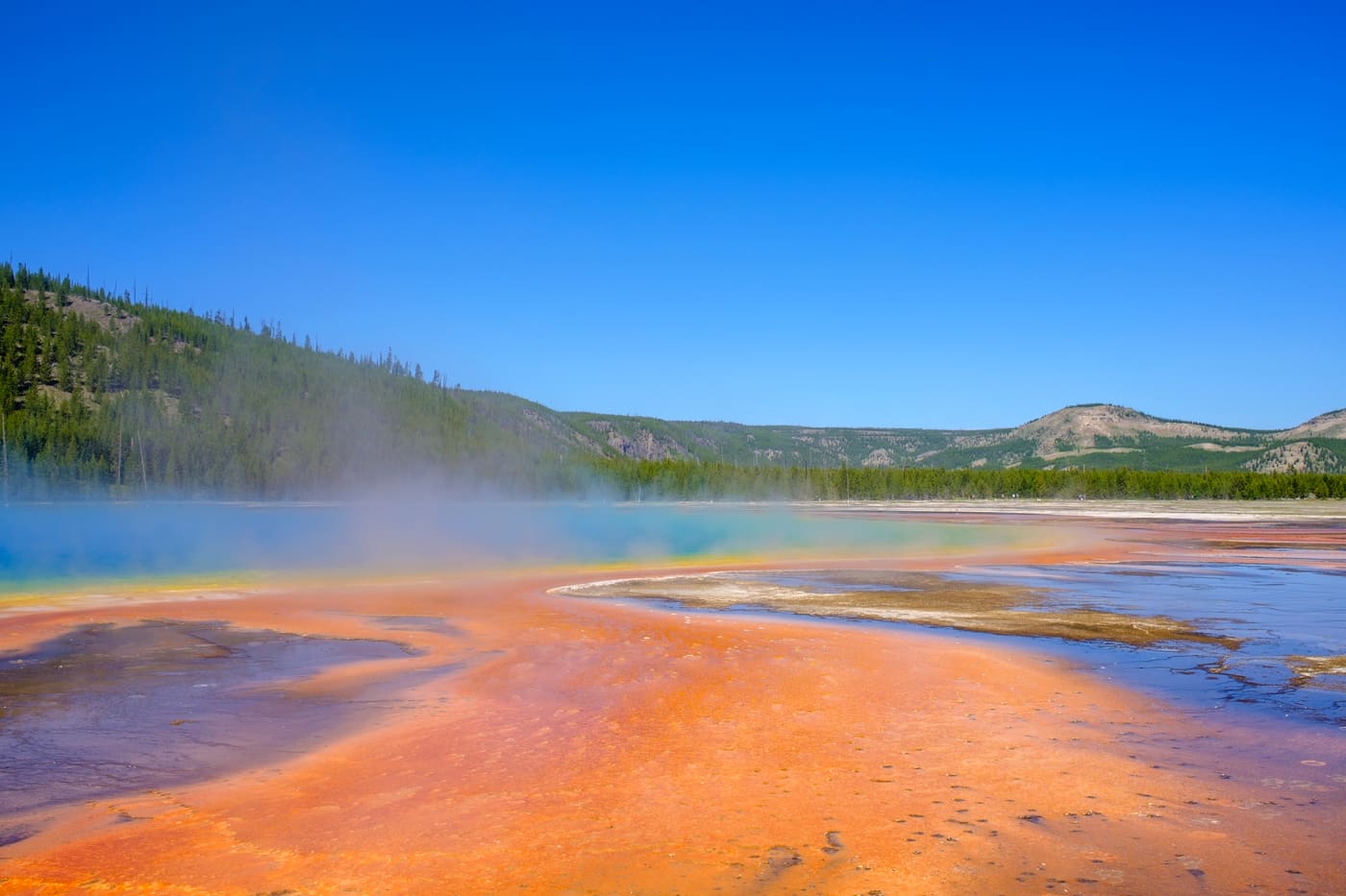 Steam drifts over an orange and turquoise thermal spring below forested hills under a clear blue sky.