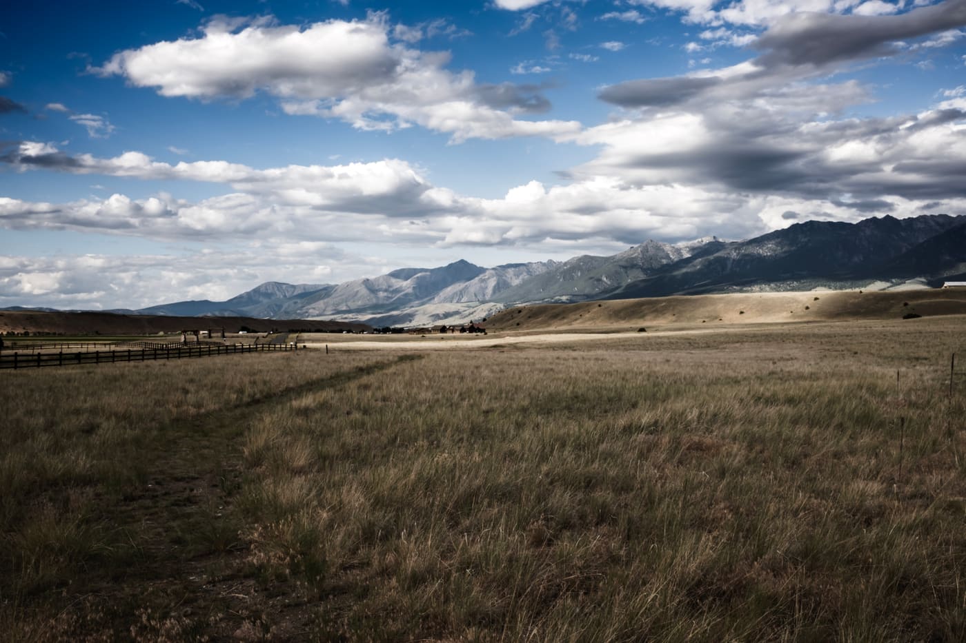 Golden grassland with a wooden fence line leading toward snow-dusted mountains under a wide cumulus-filled sky.