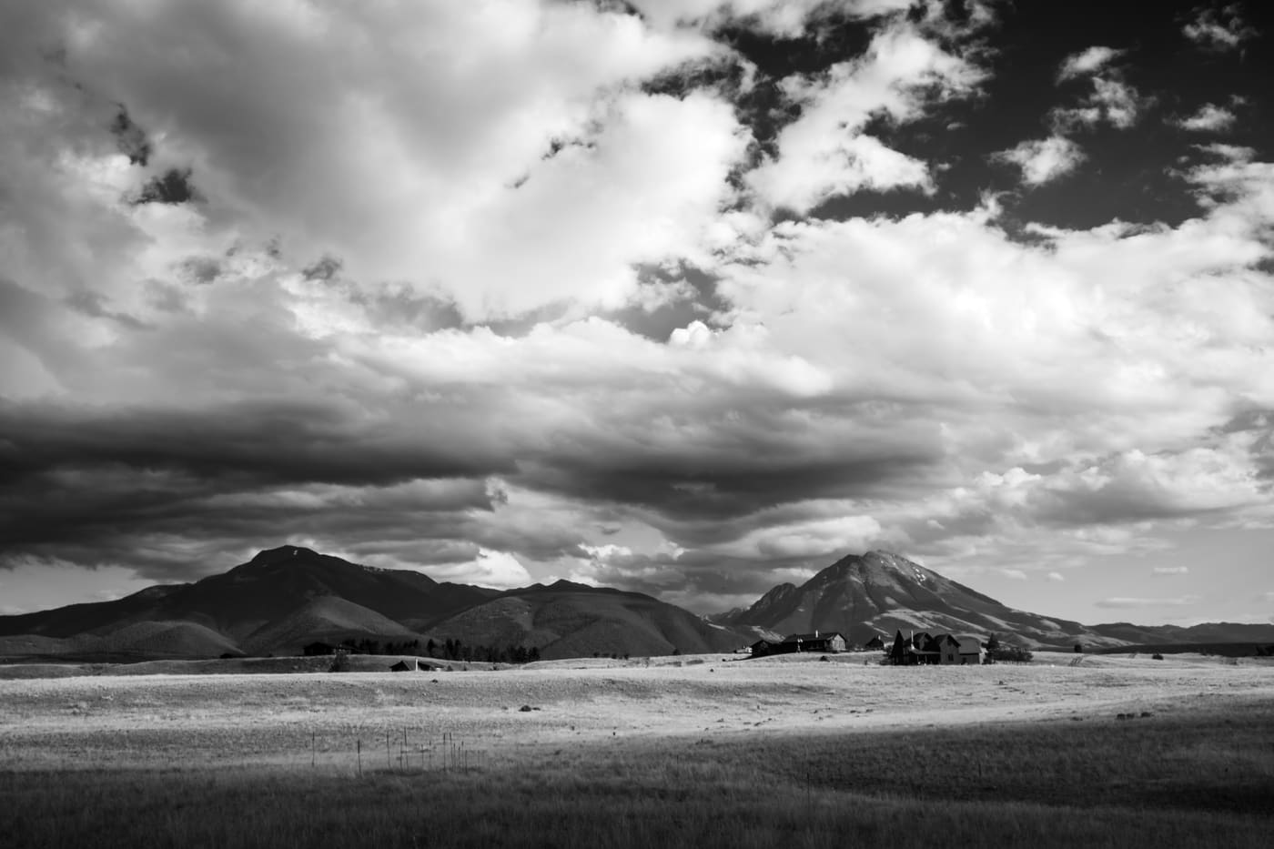 Black and white wide valley with distant ranch buildings below rolling foothills and towering cumulus clouds.