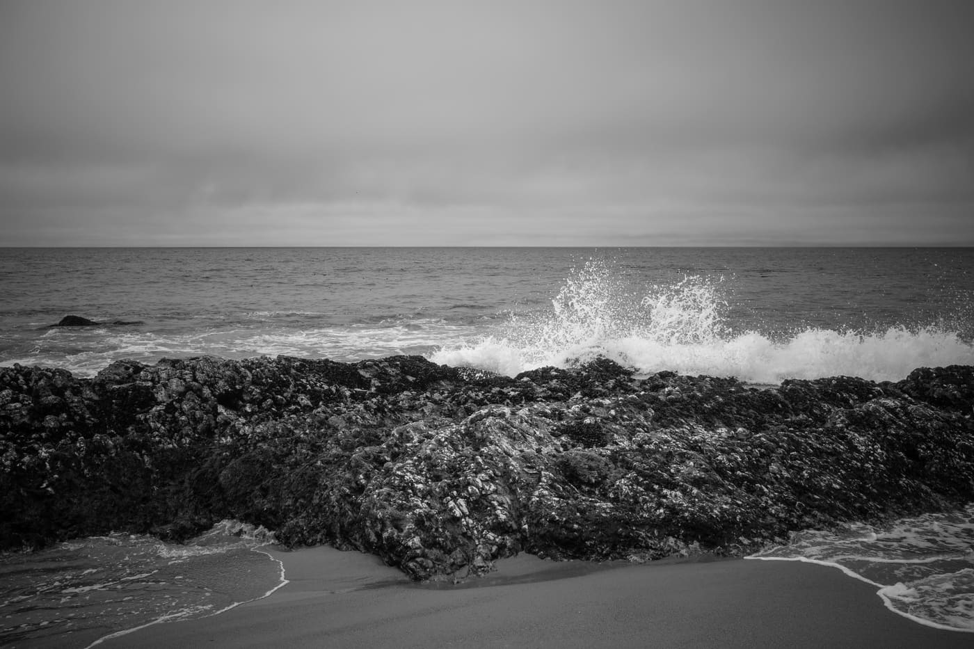 Black and white seascape of a wave bursting into spray behind a dark rocky reef, with wet sand in the foreground.