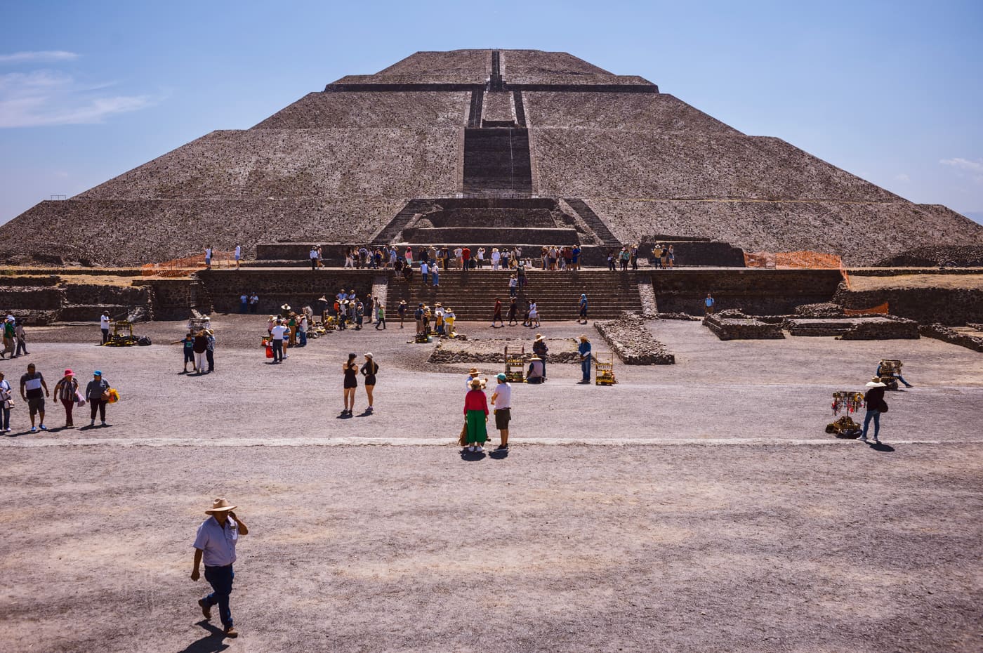 Visitors cross a sunlit plaza before a massive stepped stone pyramid under a pale blue sky.