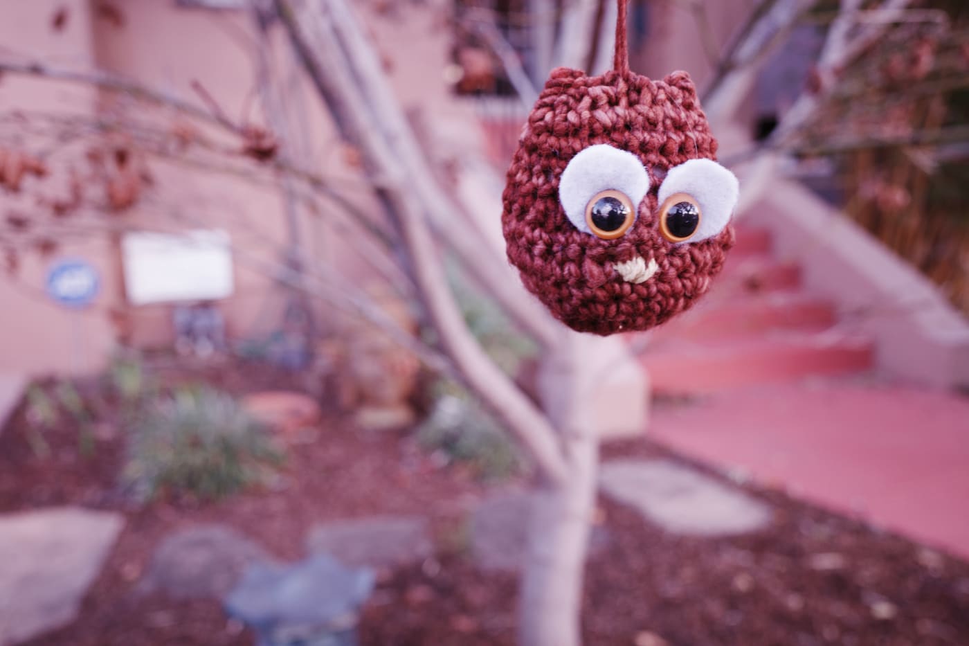 Small rust-red crocheted owl with big felt eyes hanging by a thread from a bare tree branch in a garden