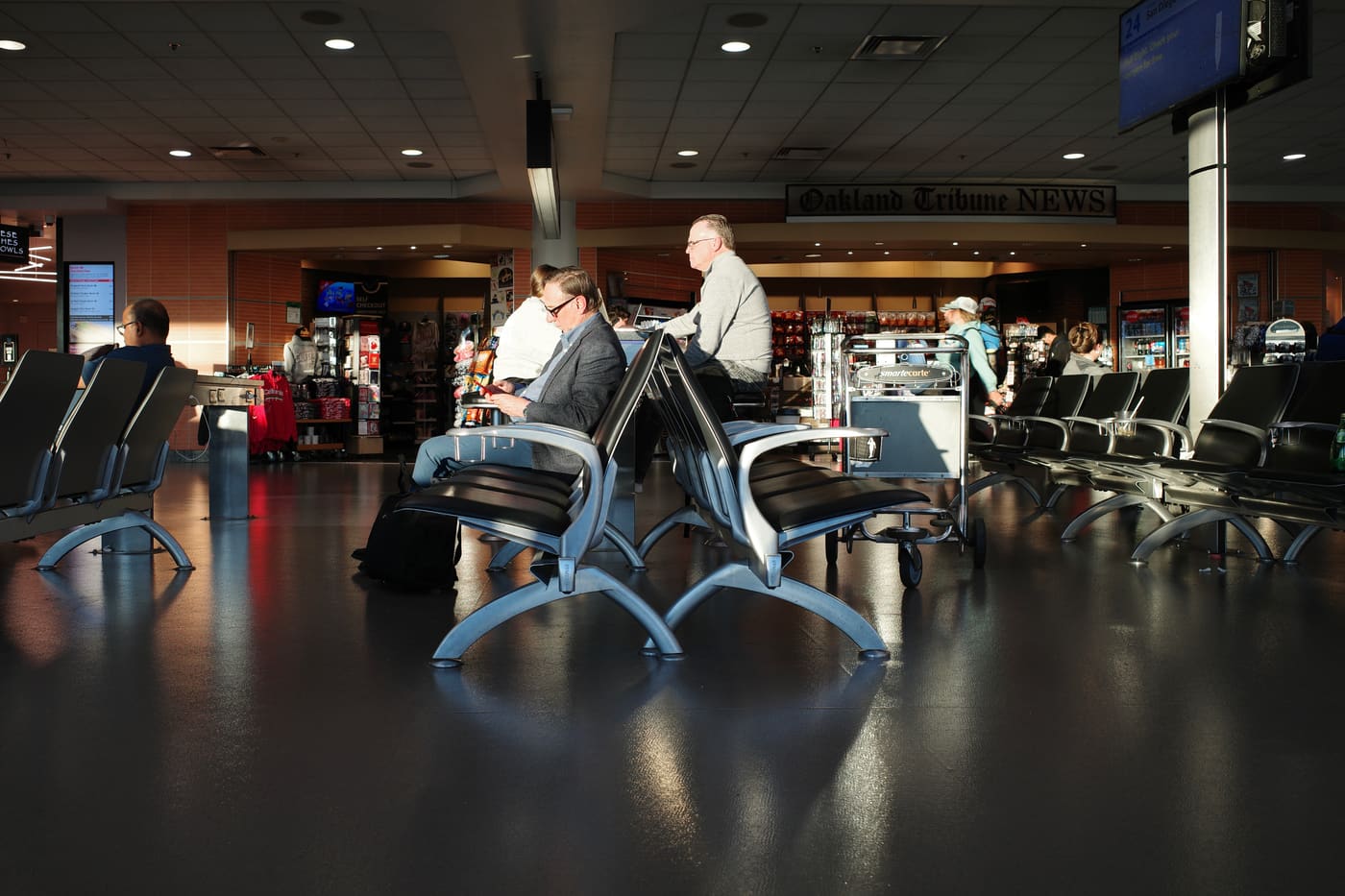 Man in grey blazer sits alone in a row of dark airport seats, sunlight raking across a polished terminal floor.