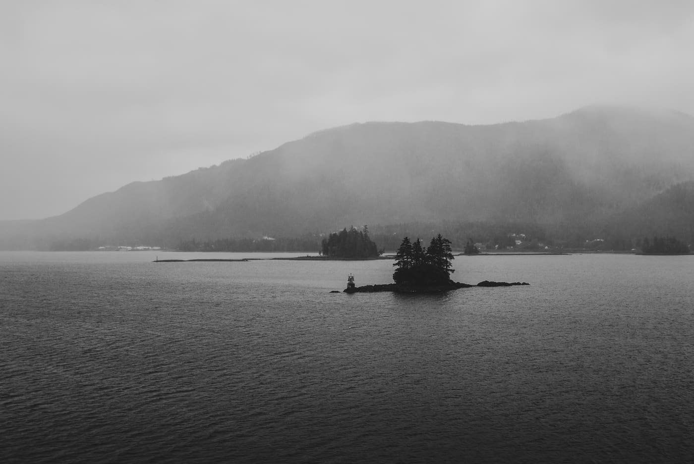 Black and white view of a tiny tree-covered island in choppy water with fog-shrouded coastal hills behind.