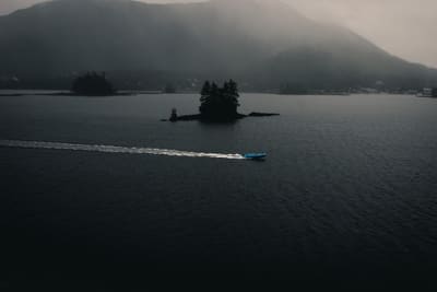 Small blue boat cutting a long white wake across dark water below fog-shrouded forested hills and a tiny island