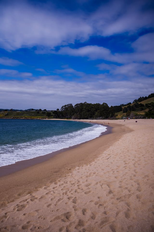 Wide sweep of pale sand curving beside turquoise surf under a dramatic blue sky streaked with white cloud.