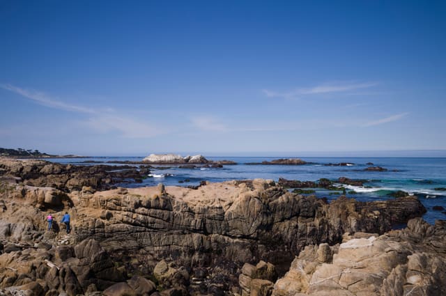 Two small hikers in pink and blue on a wide sandstone shelf above a rocky Pacific shoreline under deep blue sky.