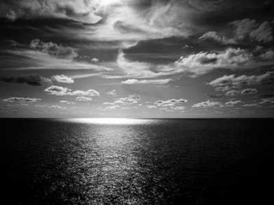 Black and white seascape with sun glare on the horizon casting a bright path across rippled open ocean under scattered clouds.