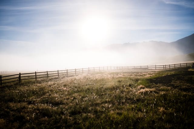 Sun burning through low fog above a wooden post-and-rail fence bordering a dry grass field with distant hills.