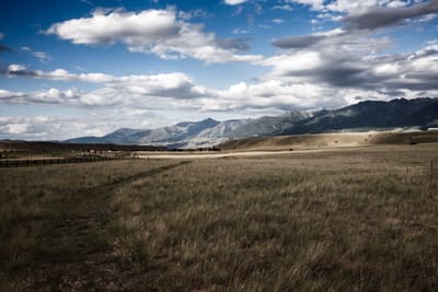 Golden grassland with a wooden fence line leading toward snow-dusted mountains under a wide cumulus-filled sky.