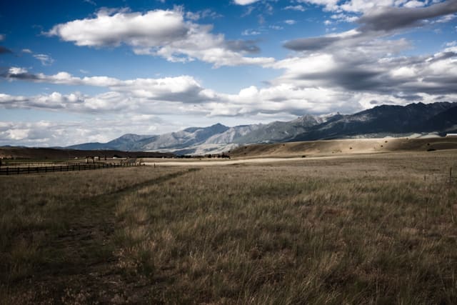 Golden grassland with a wooden fence line leading toward snow-dusted mountains under a wide cumulus-filled sky.