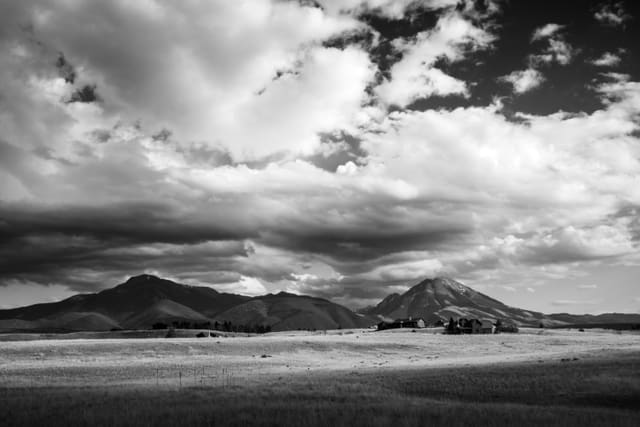 Black and white wide valley with distant ranch buildings below rolling foothills and towering cumulus clouds.