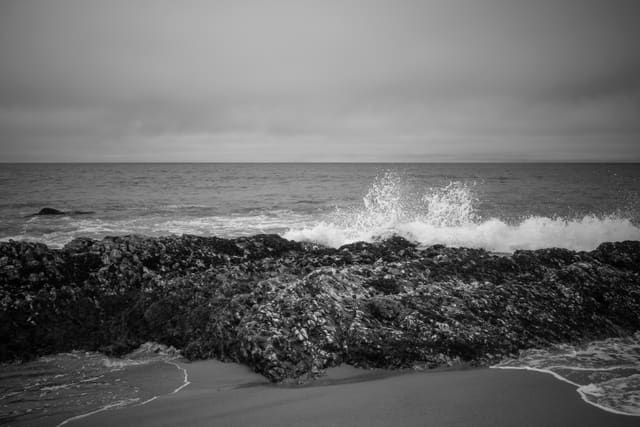 Black and white seascape of a wave bursting into spray behind a dark rocky reef, with wet sand in the foreground.