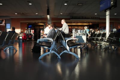 Man in grey blazer sits alone in a row of dark airport seats, sunlight raking across a polished terminal floor.