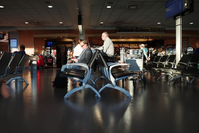 Man in grey blazer sits alone in a row of dark airport seats, sunlight raking across a polished terminal floor.